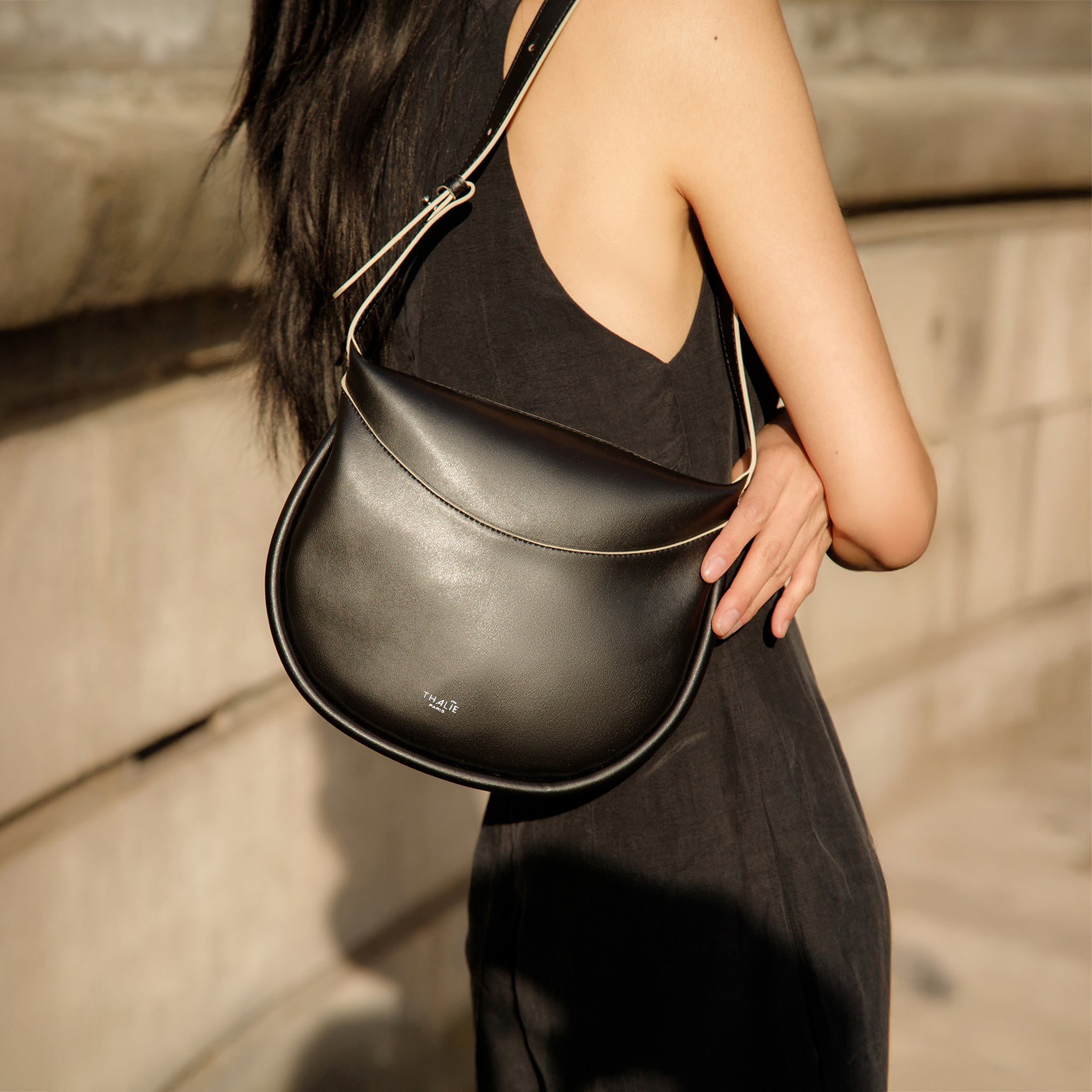 Adele black vegan cactus leather shoulder bag worn on the shoulder, featuring a sleek half-moon silhouette and minimalist design, styled with a black dress for a modern Parisian look.