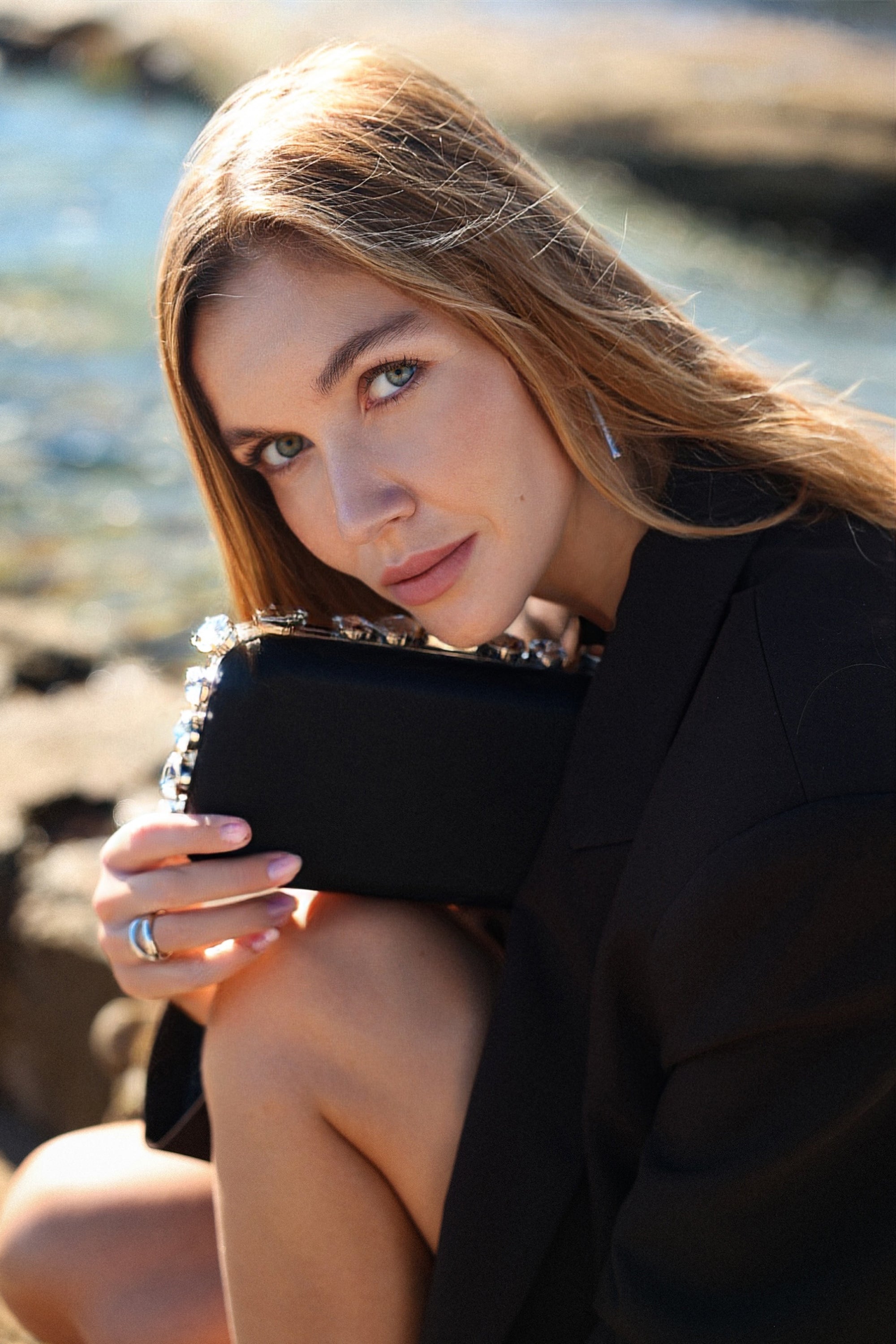 Close-up of a Thalie Paris black evening clutch with crystal detailing, styled against a sunlit seaside background.